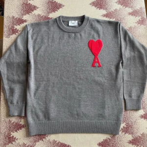 Grå tröja från Ami Alexandre Mattiussi - Säljer en stilren grå tröja från Ami Alexandre Mattiussi med ett rött hjärta och bokstaven 'A' broderat på bröstet. Tröjan har långa ärmar och ribbade muddar vid ärmslut och nederkant. Perfekt för en trendig look!