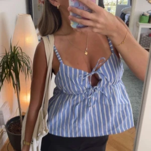 Blå- och vitrandig linnetopp - Säljer en söt blå- och vitrandig linnetopp med knytning framtill. Toppen har smala axelband och en luftig passform, perfekt för varma sommardagar. Den har en v-ringad design och är tillverkad i ett lätt och bekvämt material.