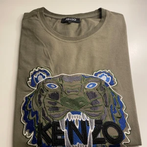 Olivgrön t-shirt från Kenzo med tigerbroderi - Snygg olivgrön t-shirt från Kenzo med ett stort, detaljerat tigerbroderi i blått, grönt och svart på framsidan. Perfekt för dig som vill sticka ut med en unik design. T-shirten har en rund halsringning och korta ärmar.