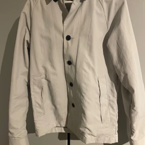 Vit jacka från Emilio - Säljer en stilren vit  jacka från Emilio med knappar framtill. Jackan har en klassisk design med långa ärmar och två fickor. Perfekt för en minimalistisk look.