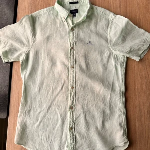 Mintrgrön linneskjorta från Gant - Snygg ljusgrön skjorta från Gant i 100% linne. Skjortan har en slim passform och korta ärmar, perfekt för varma dagar. Den har knappar framtill och en diskret broderad logga på bröstet. Tillverkad i Portugal.