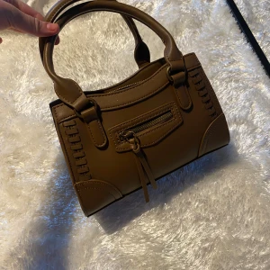 Brun handväska från Gina Tricot - Snygg brun handväska i syntetmaterial från Gina Tricot. Väskan har dubbla handtag och en praktisk dragkedja upptill. Framtill finns en dekorativ dragkedja. Aldrig använd och säljer den nu för 100!❤️