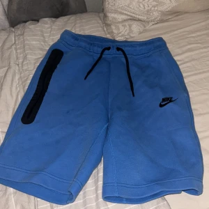 Blå shorts från Nike - Säljer ett par blå Nike shorts med svart snörning och logga. Perfekta för träning eller en avslappnad dag. De har en dragkedja på sidan för säker förvaring av småsaker.