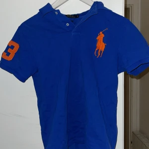 Blå pikétröja från Ralph Lauren - Snygg blå pikétröja från Ralph Lauren med orange logga på bröstet och nummerdetalj på ärmen. Mycket bra skick, storlek M. 