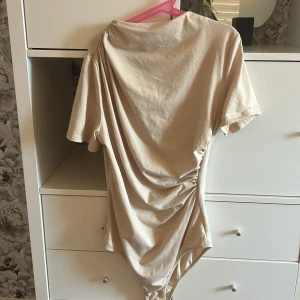 Beige kortärmad body - Säljer en stilren beige body med korta ärmar och rynkade detaljer på sidan. Aldrig använd så inga defekter. 