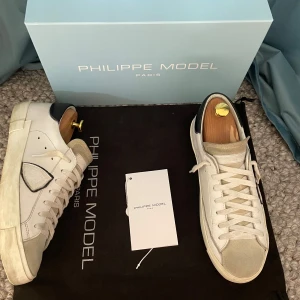 Vita sneakers från Philippe Model - Snygga vita sneakers från Philippe Model med svarta detaljer på hälen och grå mocka på tån. Skorna har en klassisk snörning och en stilren design som passar perfekt till vardags. De är tillverkade i skinn och har en bekväm passform. Nypris: 3200-3400 Pris: 2450kr Skick 8.5/10 endast en defekt (minimal lagning i högra hälen känns som normalt). Allt OG ingår förutom skoblocken.