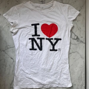 Vit t-shirt med I ❤️ NY-tryck - Säljer en klassisk vit t-shirt med det ikoniska I ❤️ NY-trycket i svart och rött. T-shirten har en rund hals och korta ärmar, perfekt för en avslappnad stil. Ett måste för alla som älskar New York! Storlek ca S