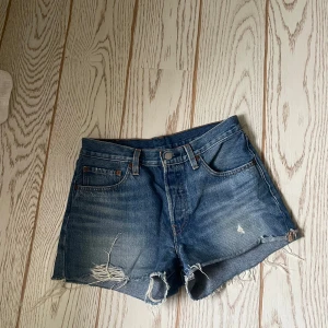 Blå jeansshorts från Levi's - Snygga blå jeansshorts från Levi's med slitna detaljer och fransiga kanter. Perfekta för en avslappnad stil. Klassisk femficksdesign och knappgylf. Har använts ca 2 gånger storlek 26 i midjan och är i mycket bra skick 