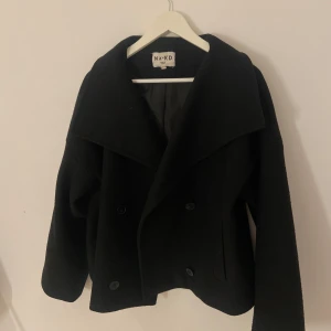 Svart kappa från NA-KD - Snygg svart kappa från NA-KD i storlek 36. Den har en klassisk design med bred krage och knappar framtill. Perfekt för kyligare dagar och ger en stilren look. Kappan är använd men det är inget som är förstört💕