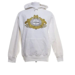 Vit hoodie från Supreme - Säljer en vit hoodie från Supreme med ett stort, detaljerat broderi i guld och svart på framsidan. Hoodien har en klassisk passform med långa ärmar och en praktisk magficka. Perfekt för dig som vill ha en stilren men ändå iögonfallande look.