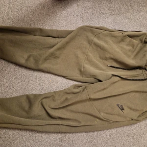 Olivgröna mjukisbyxor från Nike - Säljer ett par olivgröna mjukisbyxor från Nike med snörning i midjan. Byxorna har en avslappnad passform och är perfekta för en bekväm stil. Nike-loggan är diskret placerad på benet.