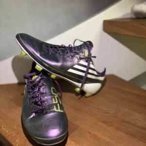 Adidas f50 - Säljer ett par lila Adidas fotbollsskor med vita och gula detaljer. Skorna har snörning och en klassisk design med dobbar för bra grepp på planen. Perfekta för fotbollsspelare som vill sticka ut.
