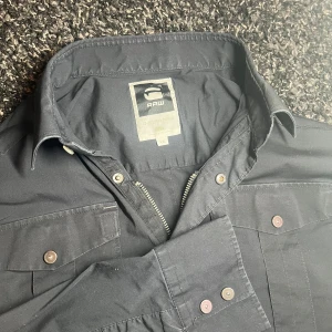 G-Star RAW overshirt - Snygg svart långärmad overshirt från G-Star RAW med knappar och dragkedja framtill. Detta är en väldigt sällsynt jacka och är väldigt eftertraktad. Skjortan har broderad text på ryggen och två bröstfickor med knappstängning. Perfekt för en stilren look. Det står att den är i XXL men jag skulle säga att den sitter som L. Den är perfekt nu till våren och sommaren!