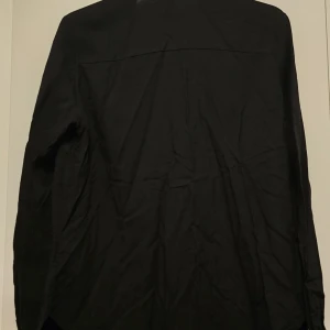 Svart skjorta från H&M Divided - Säljer en stilren svart skjorta från H&M Divided. Skjortan har långa ärmar och knappar framtill. Perfekt för en minimalistisk look. Passar bra till både jeans och chinos.