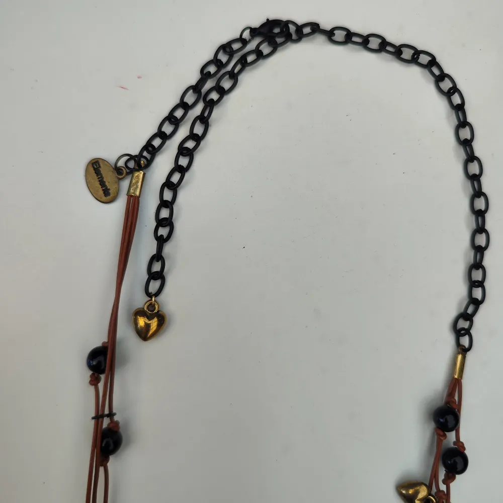 Unikt halsband med ett stort svart hjärta som hänge. Halsbandet har flera dekorativa pärlor i rosa och guld samt ett litet guldfärgat hjärta. Kedjan är i svart och brunt läderliknande material.. Asusteet.
