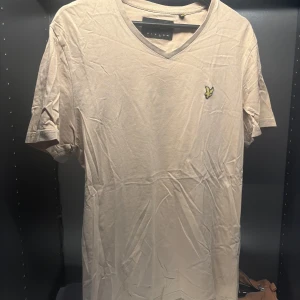 Beige t-shirt från Lyle & Scott - Säljer en stilren beige t-shirt från Lyle & Scott med v-ringning och korta ärmar. Den har en liten broderad logga på bröstet. Perfekt för en avslappnad look.