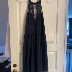 Mörkblå spetsklänning från NLY Eve - Säljer en elegant mörkblå klänning från NLY Eve med vacker spetsdetalj upptill och en luftig kjol. Klänningen har en djup v-ringning och är ärmlös, perfekt för speciella tillfällen. Öppen i ryggen.
