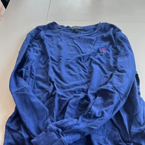Blå långärmad tröja från Polo Ralph Lauren - Säljer en snygg blå långärmad tröja från Polo Ralph Lauren med det klassiska röda logotypbroderiet på bröstet. Perfekt för en stilren och avslappnad look. Tröjan är tillverkad i mjukt bomullsmaterial.