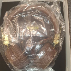Bruna trådlösa hörlurar från Guess - Snygga trådlösa hörlurar från Guess i brunt med guldiga detaljer. De har ett stilrent mönster och bekväma öronkuddar. Perfekta för musikälskare som vill ha både stil och funktion. Kvitto och laddare finns med. Ny i kartongen 