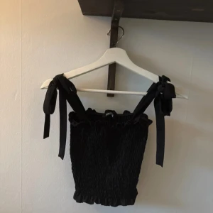 Svart topp från Gina Tricot - Snygg svart topp från Gina Tricot med knytband på axlarna. Toppen har en smockad design som ger en tight passform och volangdetaljer vid kanten. Perfekt för en stilren look.