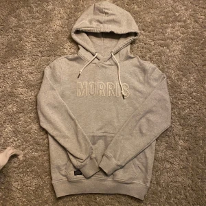 Grå hoodie från Morris - Säljer en snygg grå hoodie från Morris med broderad logga på bröstet. Den har en klassisk design med dragsko i huvan och ribbade muddar vid ärmslut och nederkant. Perfekt för en avslappnad stil. 🩶