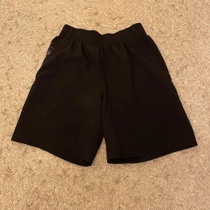 Svarta shorts från Under Armour - Säljer ett par svarta idrottsshorts från Under Armour i storlek M. De har en loose passform och är perfekta för träning. Shortsen har en elastisk midja och ett coolt tryck längs den sidan av märkesnamnet. 