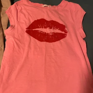 Rosa t-shirt med rött läpptryck från Coster Copenhagen - Säljer en rosa t-shirt från Coster Copenhagen med ett stort rött läpptryck på framsidan. T-shirten har korta ärmar och en rund halsringning. Perfekt för en trendig och avslappnad stil. Skriv om ni har prisförstag eller frågor☺️☺️