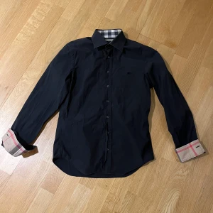 Burberry skjorta - Snygg svart skjorta med långa ärmar och klassisk krage. Den har stilrena knappar och unika rutiga detaljer på insidan av kragen och ärmsluten. Perfekt för en elegant look.