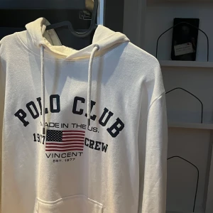 Vit hoodie från Polo Club - Säljer en stilren vit hoodie från Polo Club med tryck på bröstet och ett broderat emblem på ärmen. Hoodien har en klassisk design med dragsko i huvan och en stor ficka framtill. Perfekt för en avslappnad stil.