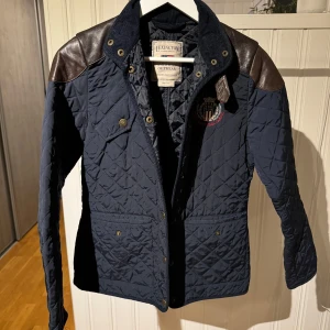 Mörkblå quiltad jacka från Lexington - Säljer en snygg mörkblå quiltad jacka från Lexington med bruna skinnaxlar och detaljer. Jackan har knappar och en broderad logga på bröstet. Perfekt för en stilren look.