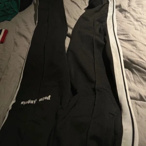 Svarta mjukisbyxor med vit rand - Säljer ett par svarta mjukisbyxor med en vit rand längs sidorna. Byxorna har texten 'Palm Angels' tryckt på ena benet. Perfekta för en avslappnad stil.