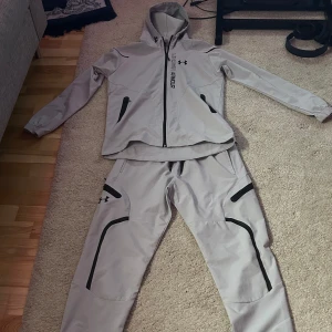 Grå träningsjacka från Under Armour - Säljer en grå träningsjacka från Under Armour med dragkedja och huva. Jackan har långa ärmar och svarta detaljer, inklusive logotyp och dragkedjor på fickorna. Perfekt för sportiga aktiviteter.