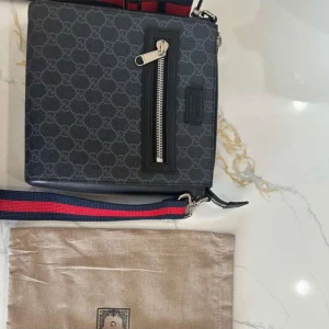 Svart axelväska från Gucci - Snygg svart axelväska från Gucci med ett subtilt mönster och en praktisk dragkedja framtill. Väskan har en justerbar axelrem i rött och blått för extra stilpoäng. Perfekt för att bära dina nödvändigheter med stil.