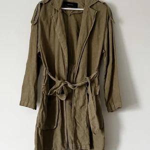 Grön trenchcoat med knytband - Snygg grön trenchcoat i linne. Jackan har långa ärmar och ett knytband i midjan för en justerbar passform. Den har dekorativa detaljer på axlarna och är perfekt för vår och höst. Storlek XS (S).