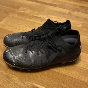 Svarta fotbollsskor från Puma - Snygga svarta fotbollsskor från Puma med vit logga på ovansidan. Skorna har snörning och en strumpliknande passform för extra komfort. Perfekta för fotbollsspel på gräsplaner.