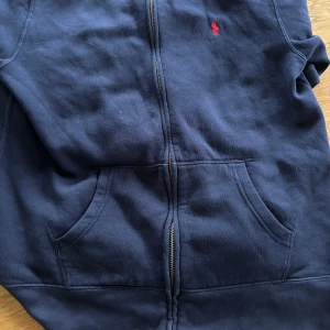 Mörkblå hoodie från Ralph Lauren - Säljer en snygg mörkblå hoodie från Ralph Lauren med dragkedja och det klassiska röda logotypen på bröstet. Tröjan har långa ärmar och en praktisk ficka framtill. Perfekt för en avslappnad stil.