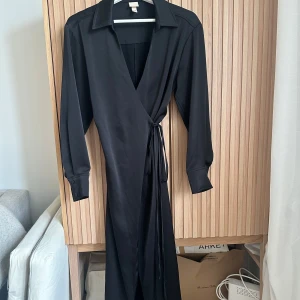 Svart omlottklänning - Elegant svart omlottklänning med långa ärmar och knytband i midjan. Klänningen har en v-ringad design och är tillverkad i ett mjukt material som ger en fin passform. Perfekt för en stilren look.