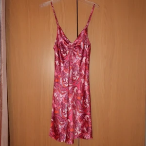 Mönstrad sidenklänning - Säljer en färgglad klänning i siden med ett livfullt mönster i rosa, rött och lila. Klänningen har smala axelband och en v-ringad design som ger en elegant touch. Perfekt för sommarens festligheter!