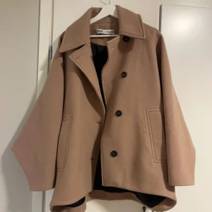 Beige kappa från Zara - Säljer en stilren beige kappa från Zara med stora svarta knappar. Jackan har en klassisk krage och långa ärmar, perfekt för höst och vår. Den har en lös passform som ger en avslappnad look.