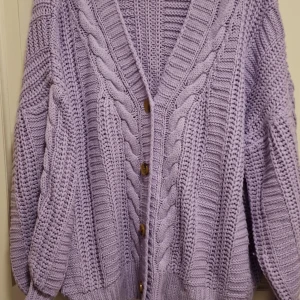 Lila stickad kofta med knappar - Mysig lila stickad kofta med ett flätmönster och träknappar framtill. Perfekt för kyliga dagar med sin avslappnade passform och långa ärmar. Ett måste för garderoben!