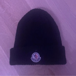 Svart stickad mössa från Moncler - Snygg svart stickad mössa från Moncler med uppvikt kant och broderad logotyp framtill. Perfekt för kyliga dagar. Äkta såklart tyvärr inget kvitto då jag köpte den begagnad. Säljer den för att den inte riktigt passa mig så bra.