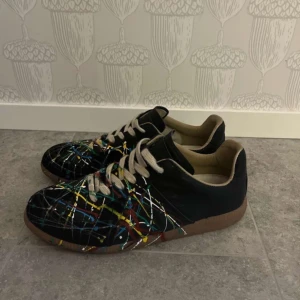 Maison margiela gats - Unika sneakers med ett färgglatt stänkmönster i blått, gult, rött och grönt på svart bas. Skorna har snörning och en brun sula. Perfekta för att sätta färg på din stil!