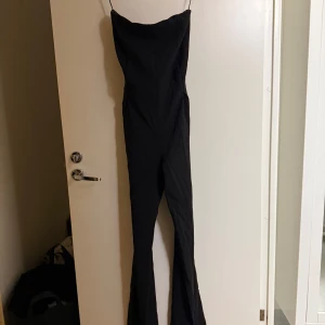 Svart axelbandslös byxdress - Elegant svart byxdress med axelbandslös design och smala axelband. Klänningen har en stilren passform och en dekorativ knytning i midjan. Perfekt för en kväll ute eller en speciell tillställning.