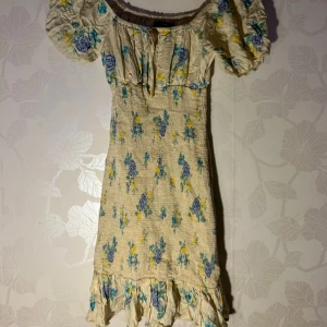 Blommig klänning från Bikbok - Charmig blommig klänning från Bikbok i beige med blå och gula blommor. Klänningen har en offshoulder-design med puffiga ärmar och volangdetaljer vid fållen. Perfekt för en somrig look!
