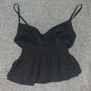 Svart ärmlös topp - Snygg svart ärmlös topp med smala axelband och v-ringad design. Toppen har en elastisk midja som ger en fin passform och en lätt volangdetalj nedtill.