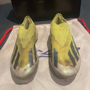Adidas Crazyfast elite Fg  - Säljer ett par gula Adidas fotbollsskor med blå detaljer. Skorna har en strömlinjeformad design utan snörning och är perfekta för snabba rörelser på planen. De har en lätt och flexibel konstruktion.
