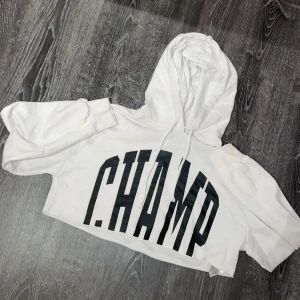 Vit croppad hoodie från FB Sister - Säljer en vit croppad hoodie från FB Sister med stor text 'CHAMP' i svart på framsidan. Perfekt för en avslappnad stil med en modern twist. Hoodien har en justerbar huva och korta ärmar. Den är från FB sister men köpt på new yorker!