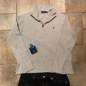 Ralph Lauren Quarter Zip Medium - Säljer en stilren grå Quarter/Half Zip trja från Polo Ralph Lauren med dragkedja vid halsen och det klassiska logotypbroderiet på bröstet. Perfekt för en avslappnad och snygg look. Riktigt fint skick.