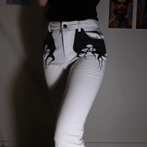 Vita byxor med svart flammönster - Snygga vita byxor med ett coolt svart flammönster på framsidan och vid bensluten. Byxorna har en hög midja, en rak passform och lätt flare. Storlek 36
