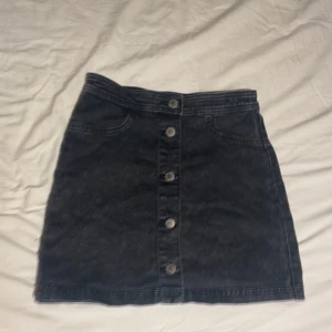 Svart jeanskjol från H&M - Snygg svart jeanskjol från H&M med knappar framtill. Perfekt för en stilren look. Kjolen har en klassisk design med fickor och är lätt att matcha med olika toppar.